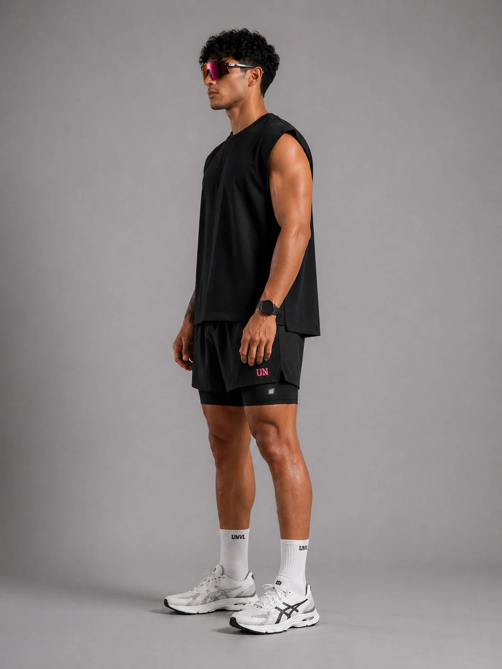 Man wearing a black sleeveless shirt and shorts with white sneakers on a gray background